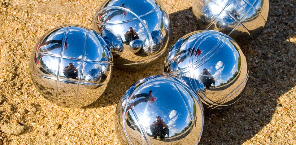 Fünf glänzende Boulekugeln liegen auf sandigem Untergrund, reflektieren Licht und Umgebung.